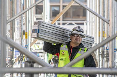 A man wearing a construction vest carries four metal beams on a construction site.