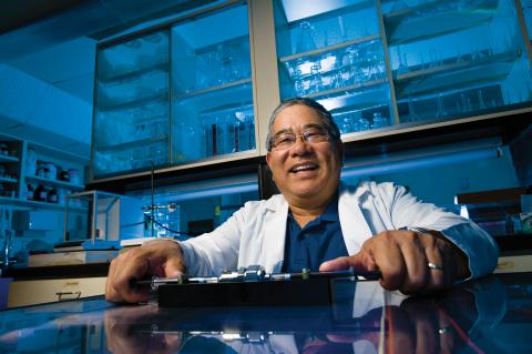 Terry Matsunaga in a laboratory lit with blue light, wearing a lab coat and smiling.