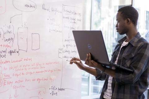 A student holds a laptop and writes on a dry erase board.