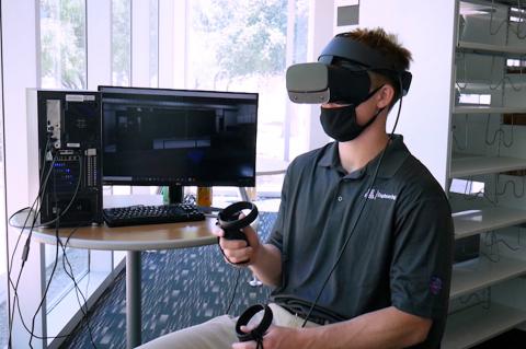 A student sitting next to a computer monitor and wearing a VR headset.