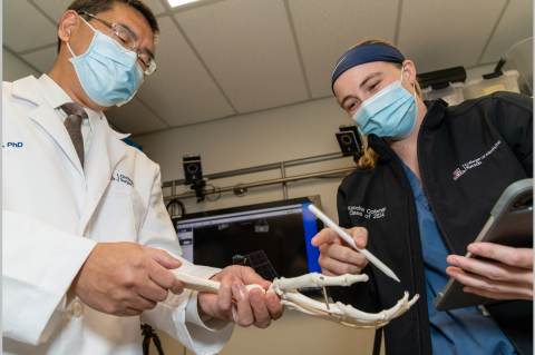 A professor and a student, both wearing surgical masks, seen from below. They are looking down at a model of the hand and wrist bones.