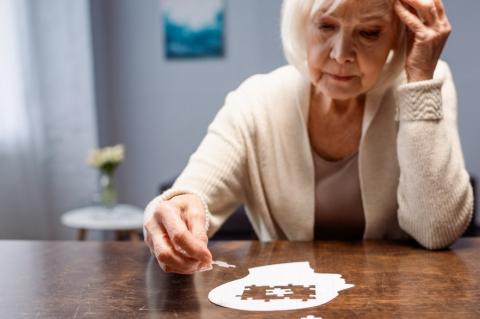 An older woman sitting at a table completing a white puzzle shaped like a human head.