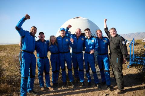 A group of eight people in blue jumpsuits smile in front of a round spacecraft. They look happy, with their arms around each other or raised in the air in celebration.