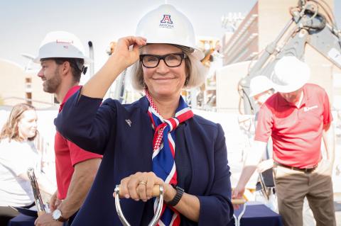 Betsy Cntwell on a construction site, wearing a white hard hat with a Block A logo. She is smiling and holding the bill of the hat.