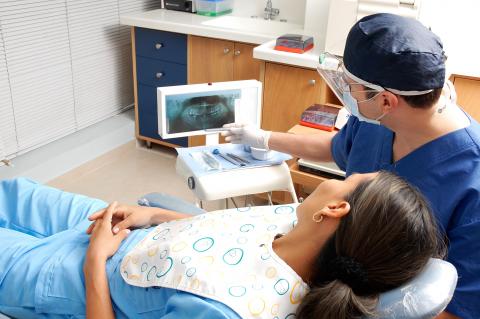 A dental patient lies in a chair while a dentist gestures toward X-rays of the patient's teeth.