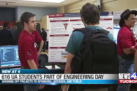 A student in a red polo stands in front of a scientific poster talking to someone in a green shirt reading the poster.