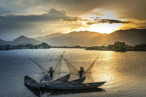 two fishers standing in water and holding up nets. There are four canoes behind them. In the distance is a sunset over mountains.