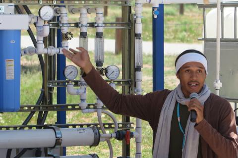 A young man holding a microphone gestures toward a systems of pipes and gauges.