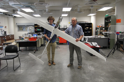 Two men stand in a large indoor space with concrete floors, holding a white sailplane about 7 feet long and with a 12 foot wingspan.