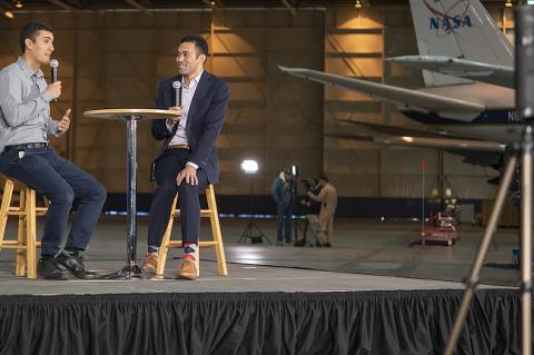 Two men sit on a stage with a NASA plane in the background