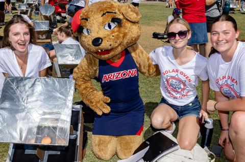 Three students and the Wilma Wildcat mascot pose outdoors with a solar oven