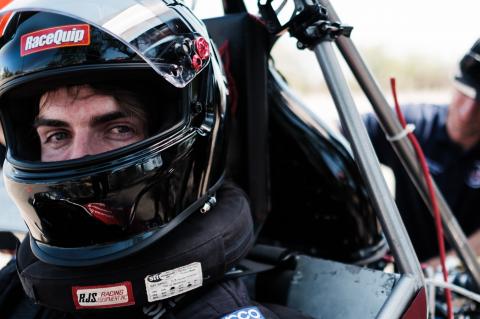 A close up photo of a student wearing a dark motorcycle helmet and sitting in a Formula-style vehicle. A man is behind him working on the vehicle.