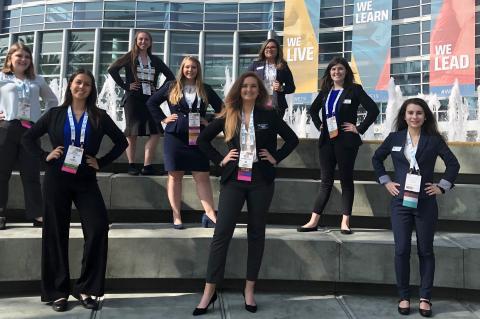 Eight women in professional attire stand on three levels of steps posing for a photo in front of the Anaheim Convention Center.