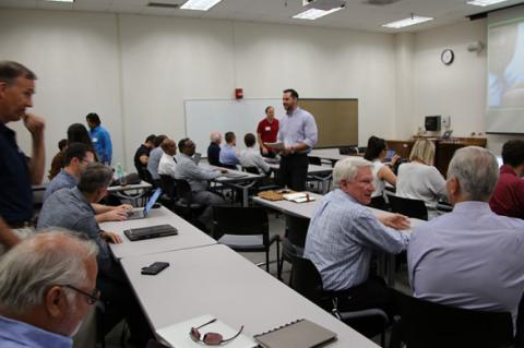 Photo taken from the back of a classroom, where students of different ages are seated and talking at long tables.