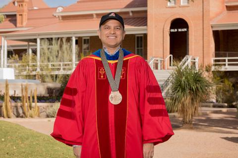 Ricardo Valerdi, wearing academic regalia and a medal, poses in front of Old Main on the University of Arizona campus 