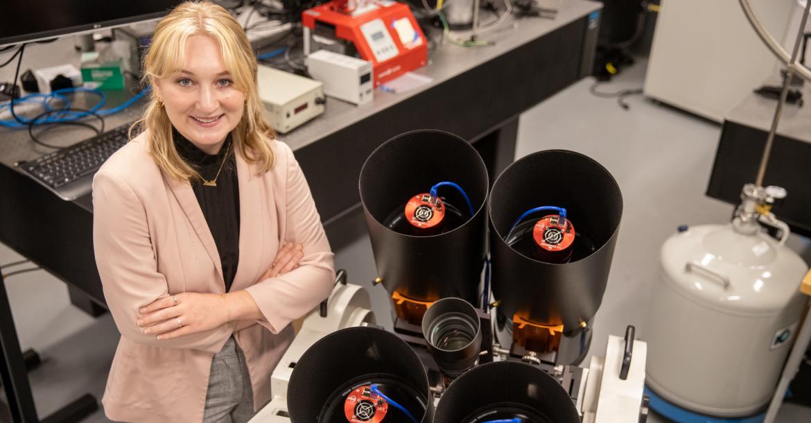 A young woman wearing a blazer is crossing her arms and smiling. She's standing in a lab environment, and next to her are four black cylinders, approximately 10 inches in diameter, win imaging instrumentation inside.