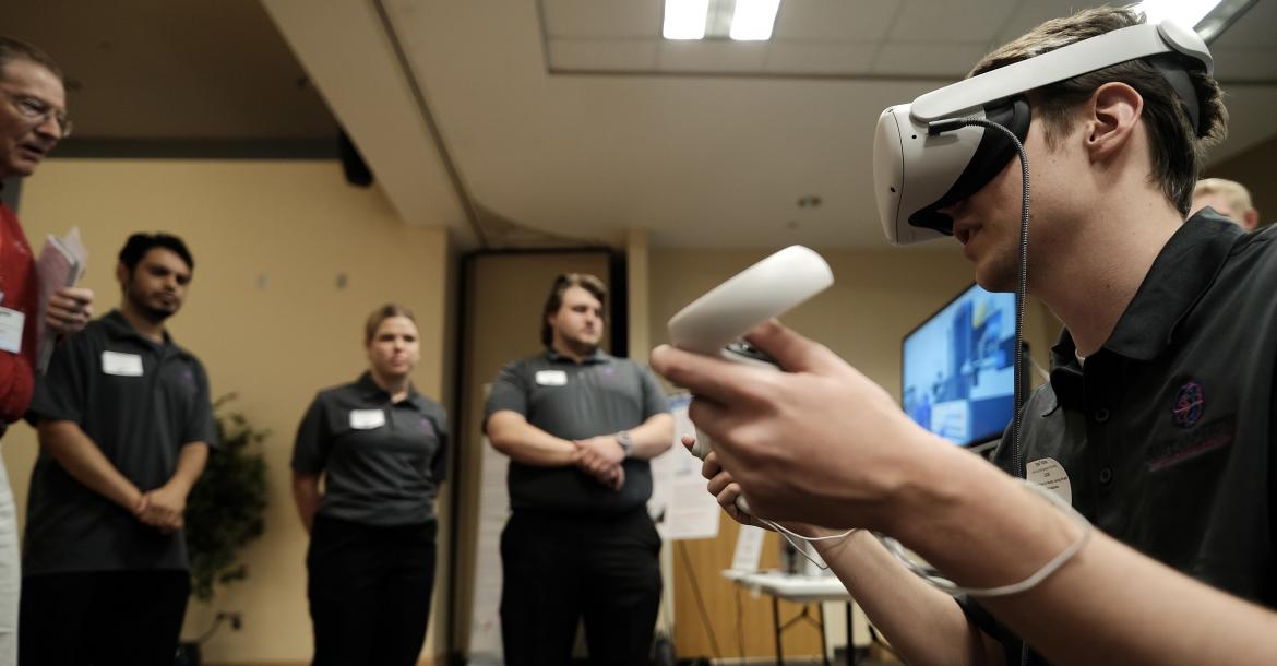 A student seen from the side wearing a VR headset and holding VR hand controllers, while students in the background look on.
