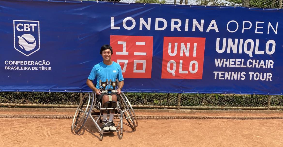Jason Keatseangsilp sits in a sports wheelchair with slanted wheels, smiling and holding two trophies on his lap. Behind him is a blue banner that reads, "Londrina Open UNIQLO Wheelchair Tennis Tour"