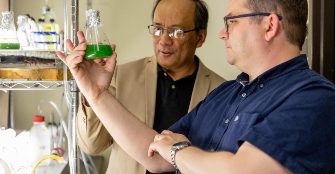 Two men examine a beaker of algae.