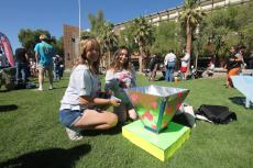 A woman with blond hair and a woman with brown hair add tape to their handmade oven on a grassy lawn.