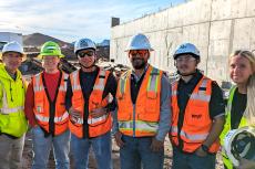 Six people pose outdoors at a construction site in hard hats