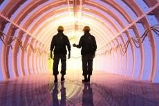Two men walking down a tunnel. They are silhouetted agains a purple-ish light coming from the end of the tunnel.