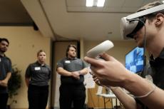A student seen from the side wearing a VR headset and holding VR hand controllers, while students in the background look on.