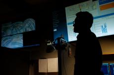 A silhouette of a man wearing glasses in front of a large computer screen.