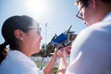 two students, seen from behind, examine a drone. the sun is shining and they are smiling.