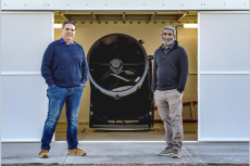 Roberto Furfaro and Vishnu Reddy stand in front of a piece of telescope equipment, a cylinder about 3-4 feet in diameter.