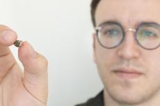 A man holds up a tiny electronic device, about the size of a chocolate chip.