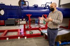 Jesse Little stands in front of a large blue wind tunnel -- it looks like a horizontal pipe -- on red scaffolding. He is wearing a black mask over his nose and mouth and gesturing with his hands to explain something.