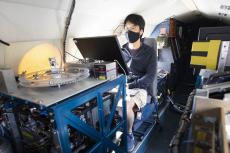 A young man wearing a face mask sits behind a monitor, surrounded by technology on a plane.