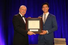 Roberto Peralta stands on a stage accepting a framed certificate from an older man. The certificate says "Astronaut Scholarship." The men are in formal wear and there is a blue curtain behind them.