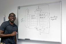 Tosiron Adegbija stands at a whiteboard with a diagram of computer memory caching written on it in black marker