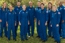 A group of ten astronauts stand in matching blue jumpsuits in a green outdoor area.