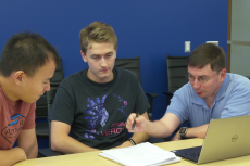 Three men lean over a laptop and set of papers on a table.