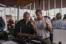 Two men stand in a crowded lobby. One is gesturing at machine on a table in front of him, while the other listens carefully.