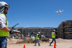 A man in a yellow construction vest and hard hat operates a drone on a construction site.