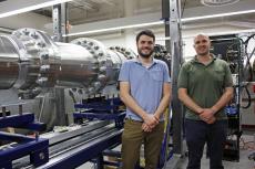 Alex Craig and Jesse Little standing next to a hypersonic wind tunnel, a horizontal metal tube about the circumference of a large tree.