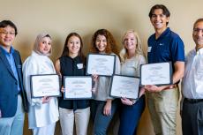 A group of seven people -- five students holding certificates flanked by two professors -- pose for a photo