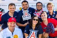 A group of seven people pose for a photo in front of a UA Engineering backdrop. They are smiling and holding props like a Block A and a foam finger in UA colors.