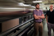 Alex Craig and Jesse Little stand next to LT5, a long wind tunnel which is silver and stretches off frame to the left of them.