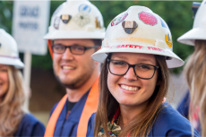 A student smiling and wearing graduation garb and a white hard hat covered in stickers. There are three other students in the frame, wearing the same combination, but out of focus.