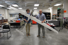 Two men stand in a large indoor space with concrete floors, holding a white sailplane about 7 feet long and with a 12 foot wingspan.