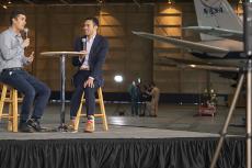 Two men sit on a stage with a NASA plane in the background