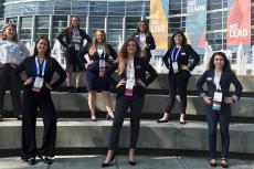 Eight women in professional attire stand on three levels of steps posing for a photo in front of the Anaheim Convention Center.