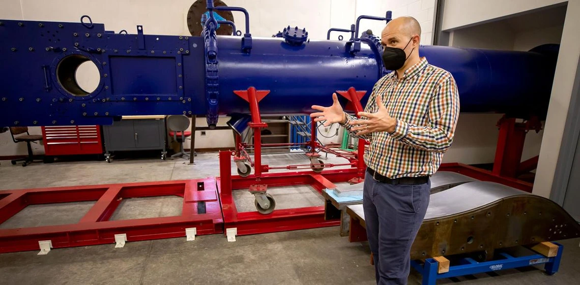 Jesse Little stands in front of a large blue wind tunnel -- it looks like a horizontal pipe -- on red scaffolding. He is wearing a black mask over his nose and mouth and gesturing with his hands to explain something.